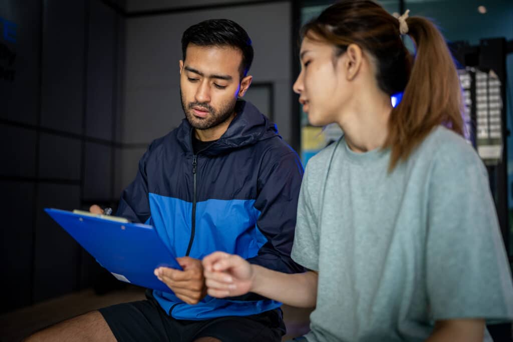 Male personal trainer holding a clipboard while discussing a workout plan with a female client in a gym.
