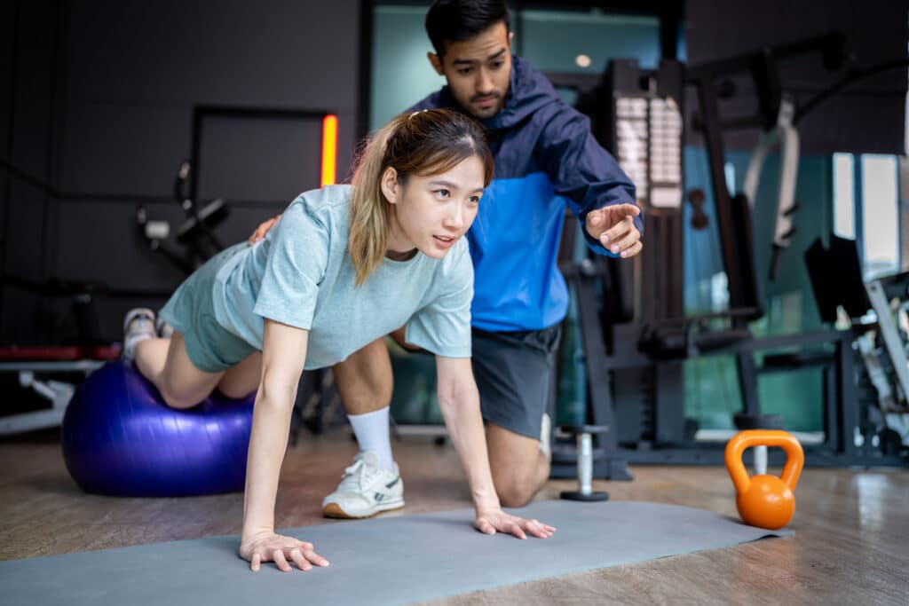 Female client performing a stability ball exercise under the guidance of a male successful personal trainer in a modern gym.