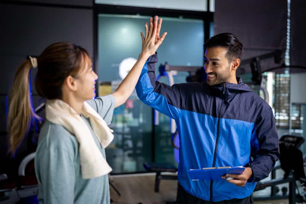 Female gym-goer high-fiving her successful personal trainer after completing a workout session, both smiling in a fitness center.