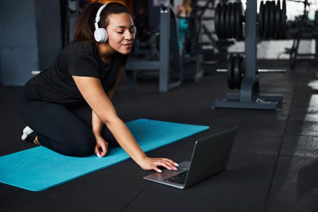 Smiling female personal trainer is sitting on mat and preparing for online training with laptop and headphones.