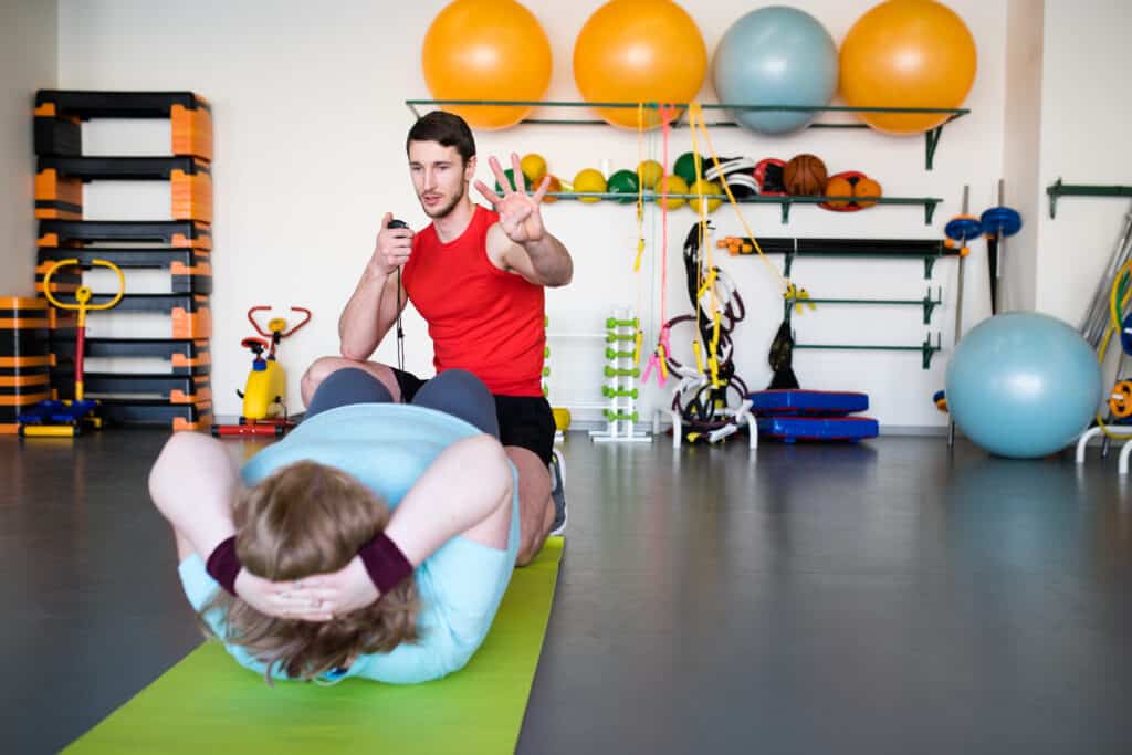 Personal trainer in a red tank top coaching a woman doing sit-ups on a green yoga mat in a gym filled with fitness equipment and colorful exercise balls.
