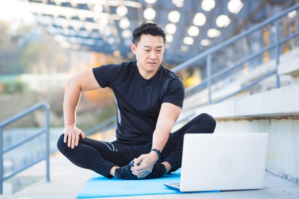 Male fitness coach holds workout classes online whilst sitting on stadium steps.