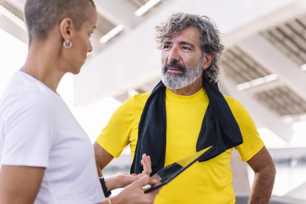Female personal trainer in a white shirt instructing a man lifting a weight plate during an exercise session in a bright gym.