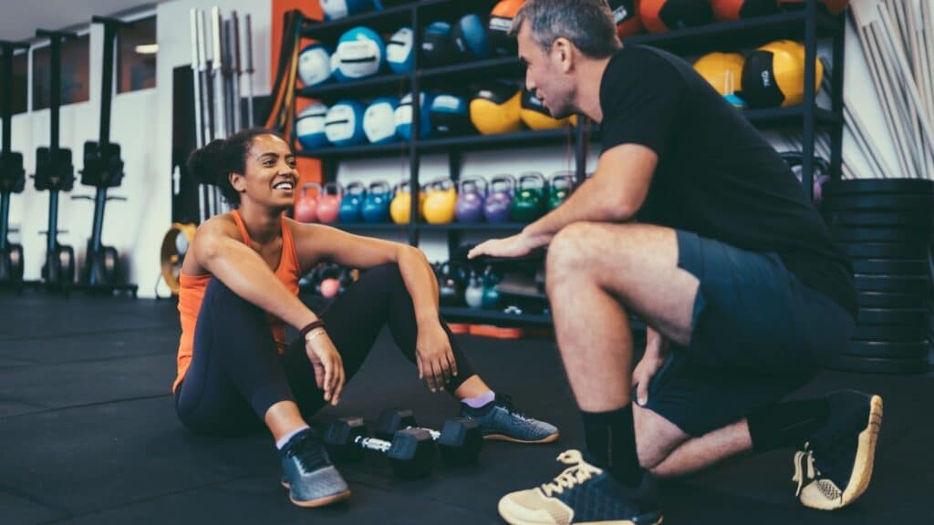 Personal trainer in conversation with a smiling female client sitting on the floor surrounded by gym equipment and weights.