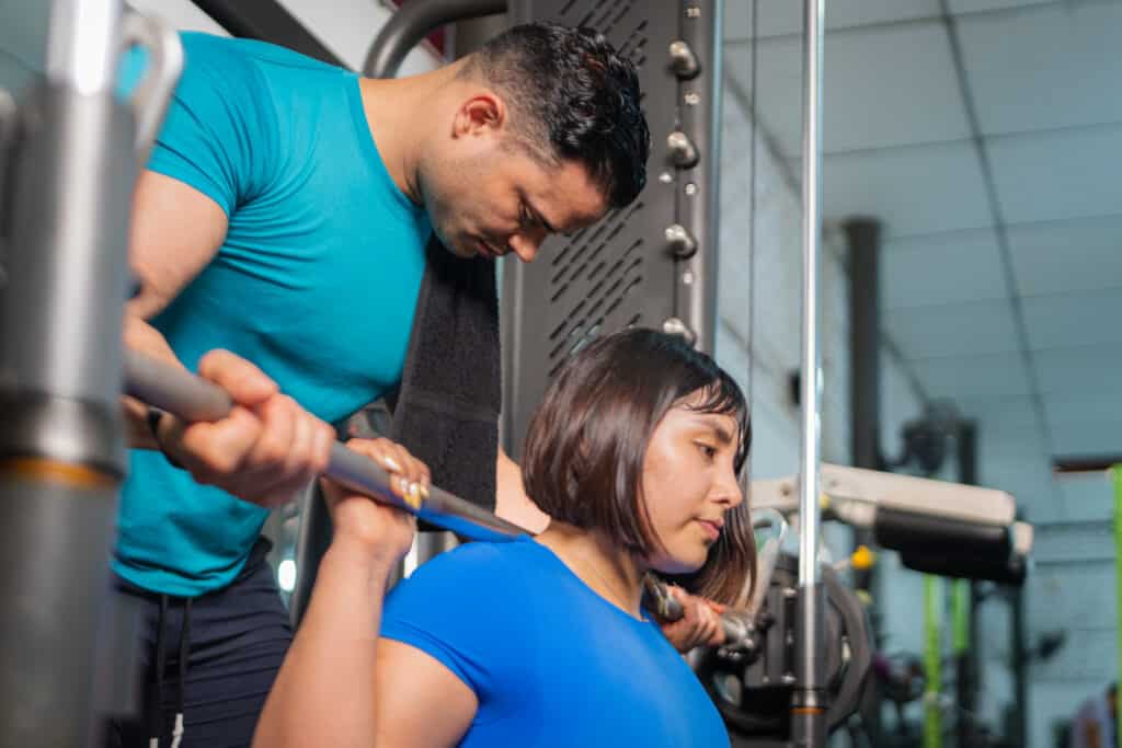 Focused woman lifting weights with personal trainer assisting and supporting her during workout session in gym.