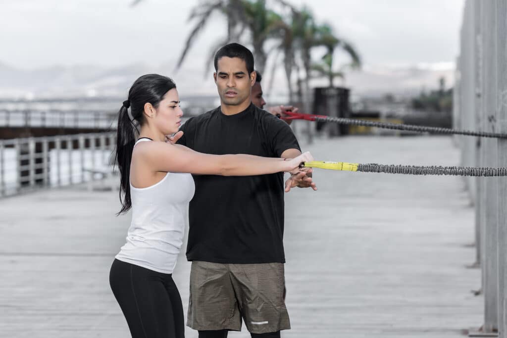 Trainer assisting a woman with a resistance band exercise on an outdoor wooden boardwalk, with palm trees and mountains in the background.
