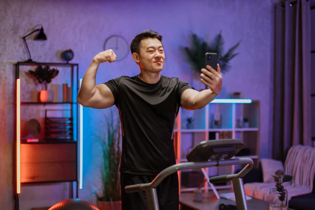 Man showing arm muscles while taking a selfie and exercising on a treadmill.