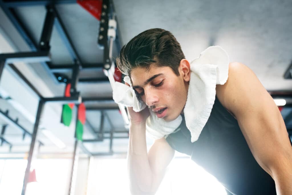 An exhausted young man in a modern gym with a white towel around his neck.